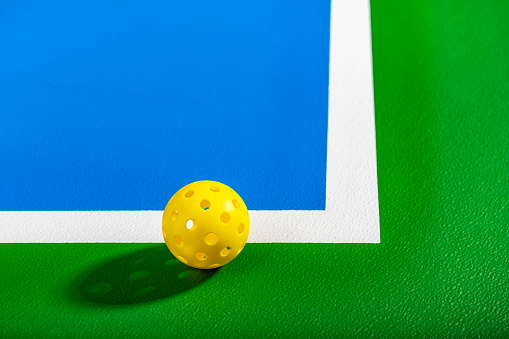 A low angle view of a yellow Pickleball ball sitting on the corner of a green and blue Pickleball court.