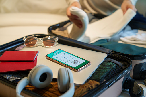 Close-up image of a smartphone displaying an electronic flight ticket next to a woman's packed suitcase containing clothing, sunglasses, headphones, a laptop, and a passport, placed on a bed. Represents the essential items for a traveler and embodies the concepts of travel, vacation, and business travel.