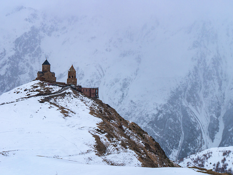 Gergeti Trinity Church sitting on top of a hill with the massive Caucasus Mountains in the background on a snowy and cold winter afternoon