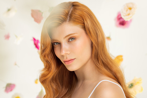 Studio shot of a beautiful redhead woman with perfect skin. Natural sunlight. Flowers floating around her.