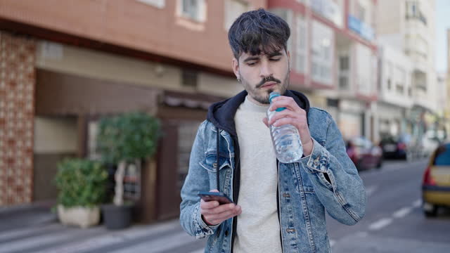 Young hispanic man standing drinking water using smartphone at coffee shop terrace