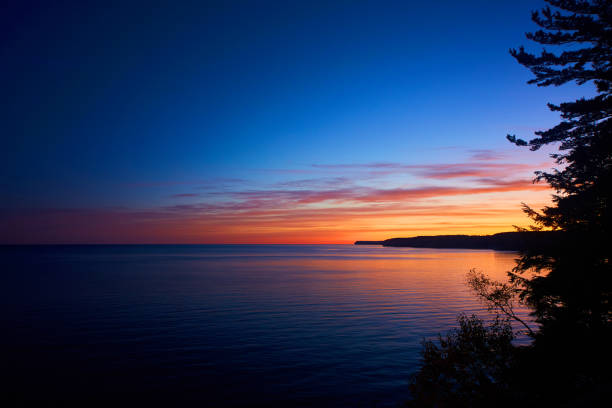 istock Endless water illuminated by an orange red and yellow sunrise framed on one side by silhouette of a pine tree and a peninsula 1490382747