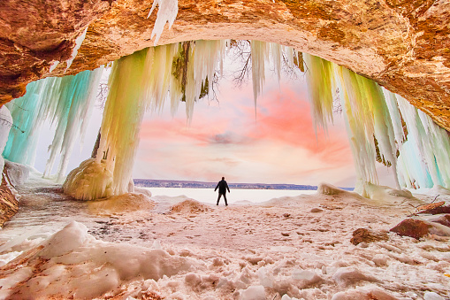 Image of Human figure silhouette standing in large ice cave entrance during winter sunrise with blue and green hanging icicles