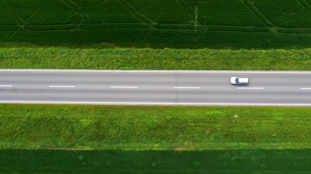 Top view drone pov aerial shot of white car driving along a straight road through cultivated landscape