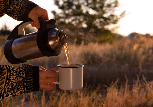 a girl pours hot tea from a thermos, autumn weather, morning tea, tea at dawn with the beautiful rays of the sun.