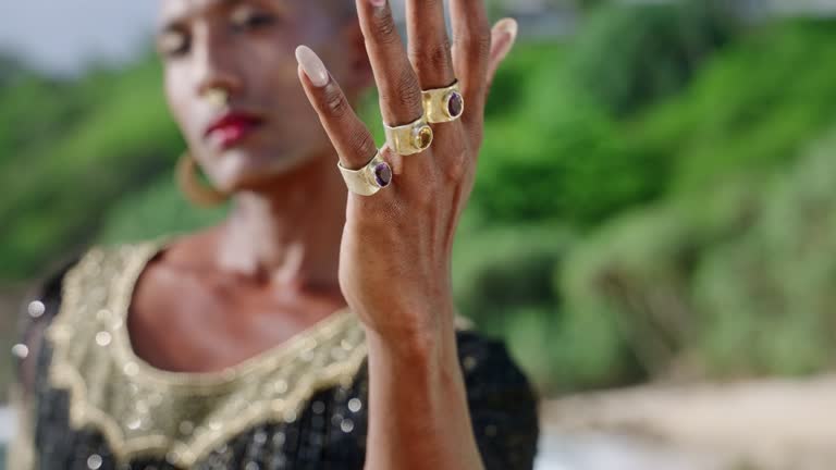 Androgynous person of color reaches out hand with luxury boutique brass jewerly on ocean beach. Ethnic fashion model wears jewellery dressed in posh gown poses in tropical location.