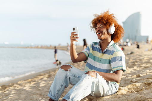 Portrait of an African American man sitting on the sandy beach and watching his favourite music videos on the smart phone