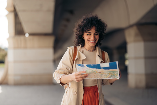 Smiling carefree young woman with curly hair in casual earth colors clothes standing outdoors sightseeing exploring in a foreign city looking for location at map.