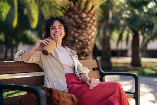 Confident happy beautiful young Italian woman looking at camera, enjoying freedom, feeling joyful and motivated, sitting outdoors in city park on sunny day, holding takeaway paper coffee cup.