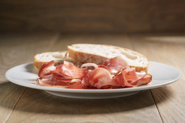 speck slices on white plate with ciabatta on old wood table, shallow focus
