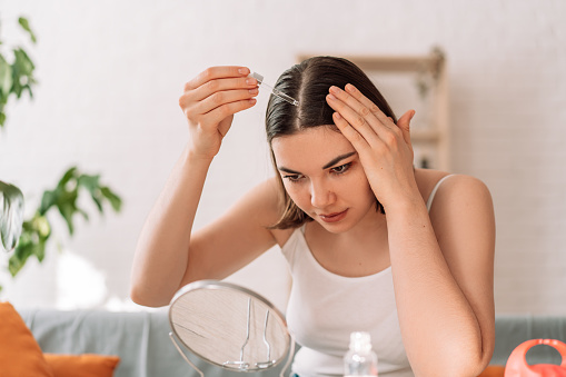 Lady sitting on the couch looks in the mirror and drips hair oil with a pipette on her hair.