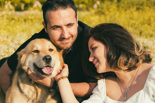 On a white picnic blanket, a pregnant woman and her husband share a tender moment, while their friendly dog seeks affection nearby