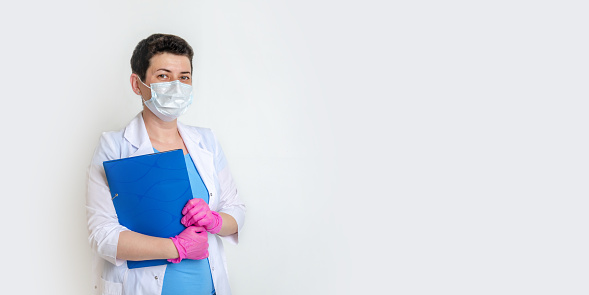 woman, doctor or nurse, dressed in medical gown with disposable mask and gloves, holds folder for documents. portrait of female doctor, trustworthy pose, looking into frame. copy space