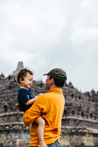 asian man carrying her son walking on the grass with Borobudur temple in the background