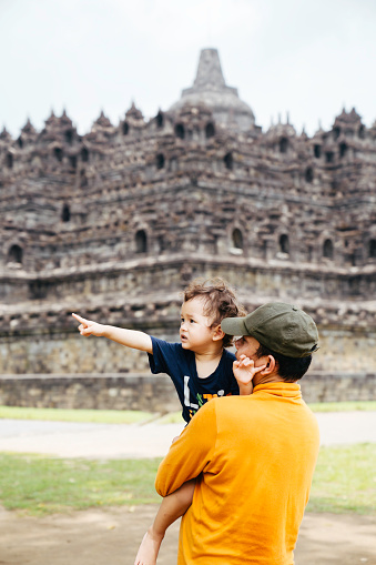 asian man carrying her son walking on the grass with Borobudur temple in the background