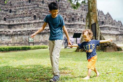 young kid with his little brother walking on the grass with Borobudur temple in the background