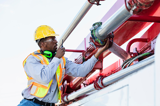 An African-American man inspecting a pipe clamp on a concrete boom pump truck, talking into a walkie-talkie. This is part of a series showing workers operating concrete pump trucks at a construction site.