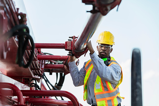 An African-American man in his 30s working as a concrete boom pump truck operator. He is climbing on the bed of the truck inspecting the boom and pipes. This is part of a series showing workers operating concrete pump trucks at a construction site.