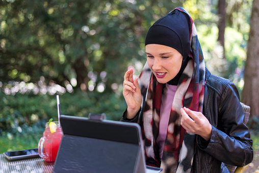 A Muslim woman in a hijab using a tablet in a street cafe next to the park.