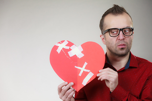 Bad relationships, breaking up, sadness emotions concept. Thoughtful adult man holding broken heart fixed with plaster bandage, on grey