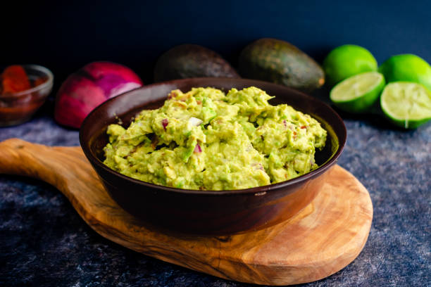 Fresh Guacamole in a Wooden Bowl with Ingredients in the Background Homemade guacamole with avocados, limes, onion, cilantro, and chipotle peppers guacamole stock pictures, royalty-free photos & images