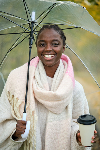 African woman with a coffee and transparent umbrella looking at camera and smiling in autumn.
