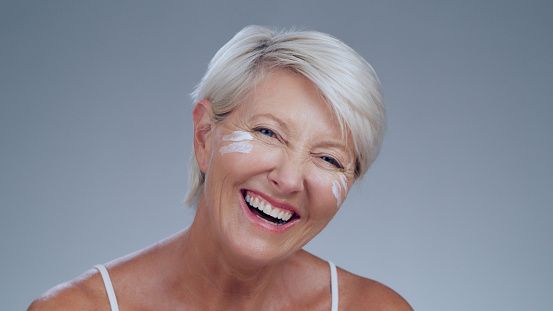 Portrait, skincare and cream with a senior woman in studio on a gray background looking happy or confident. Smile, beauty and face lotion with a mature female model feeling positive while laughing