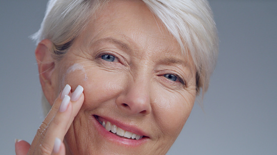 Old woman apply cream on face, beauty portrait with smile and anti aging cosmetics isolated on studio background. Moisturizer, skincare closeup and happy female person, cosmetic care and dermatology