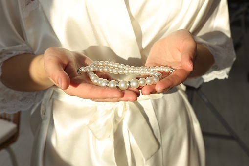 Bride holding beautiful pearl necklace indoors, closeup. Wedding day