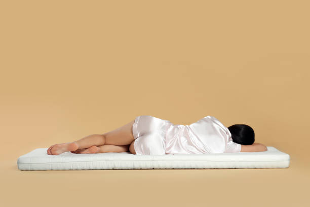 Young woman lying on soft mattress against beige background, back view