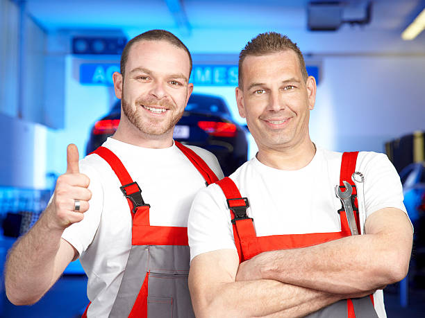 Two smiling motor mechanics are standing in a garage in front of a black car on a service lift. One of them shows thumb up.