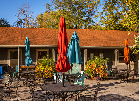 View of outdoor patio of Midwestern restaurants in small town with metal chairs and tables, colorful parasol umbrellas and potted plants; large trees behind the building