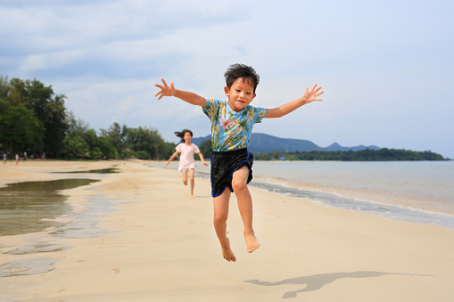 Cheerful Asian little boy child and girl kid having fun jumping and running on tropical sand beach at sunrise. Happy family sister and brother enjoy in summer holiday.
