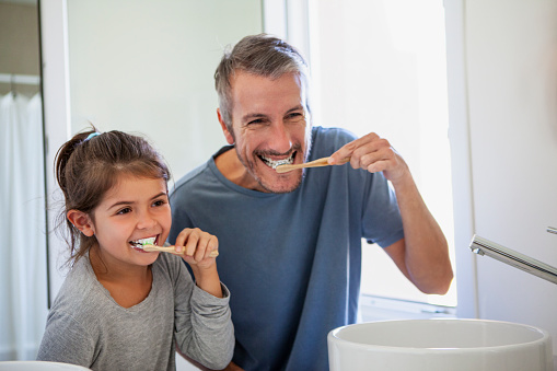 Portrait of father and daughter smiling at the mirror while brushing teeth together at home
