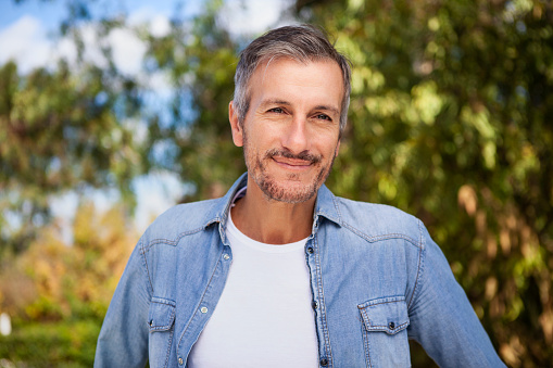 Mid-shot portrait of man with calm smile standing outdoors with trees on the background