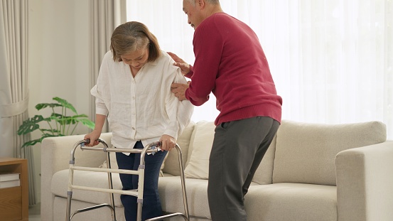 Asian senior husband take care to wife having knee pain while walking with crutch at home. Elderly asian couple giving encouragement, take care when the other is sick and doing physical therapy. Concept of family relationship, life insurance, health care, retirement.
