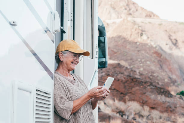 happy senior woman with eyeglasses standing outside a camper van motor home using mobile phone enjoying travel destination. smiling elderly lady in alternative vacation transport and tourism concept - motor home mobile home camping senior adult imagens e fotografias de stock