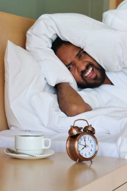 istock Image of grumpy Indian man lying in double bed with pillow covering ears, alarm clock on nightstand, bronze metal case, retro alarm clock, double bell, analogue display clock, white cup and saucer, focus on foreground 1490550047