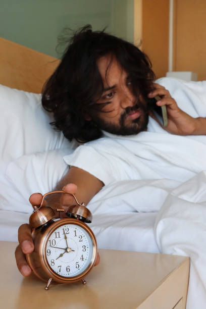 imagem em close-up do homem indiano segurando o smartphone até o ouvido, alcançando a cama de casal do hotel desligando o despertador na mesa de cabeceira, caixa de metal de bronze, despertador retrô, sino duplo, relógio de exibição analógico, foco  - double bed night table headboard bed - fotografias e filmes do acervo