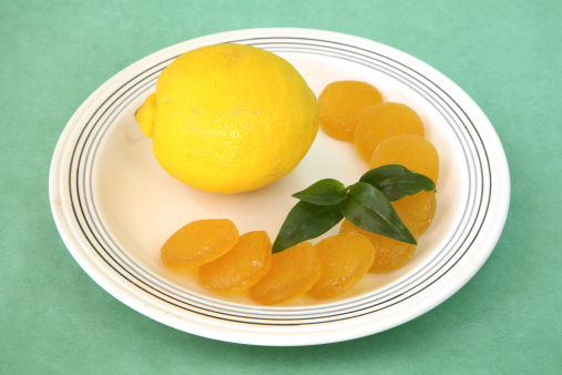 Yellow lemon on a plate with a crescent of circular lemon jelly candies in arc on right with piece of greenery in center crescent.  Light cream colored plate with thin gray lines around edge.  On plain green background.