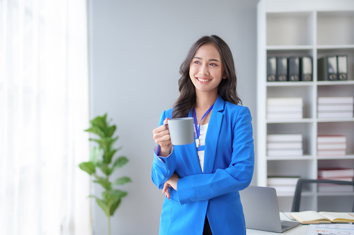 Successful Asian businesswoman smiling holding coffee cup at office. Confident Asia businesswoman standing happily in the office.