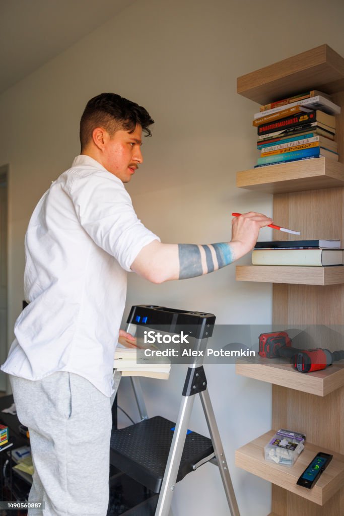 Bookshelf tidy up and keep house order. Latino man putting things in order, arranging books on the shelf, stand on stepladder in new apartment. Close up The satisfaction of man settling into a new living space and making it your own with stuff order on shelves Decluttering Stock Photo