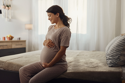 Smiling pregnant woman holding her stomach while sitting on a bed in the morning. Copy space.