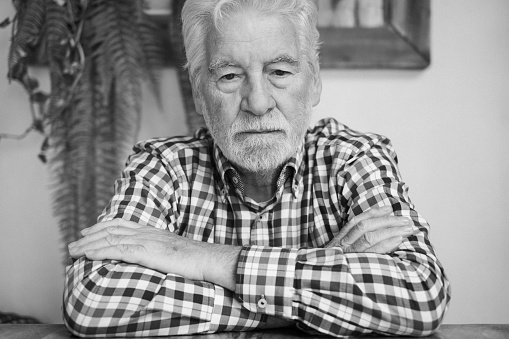 Black and white portrait of serious bearded senior man with crossed arms looking down lost in his thought while sitting at cafe table.