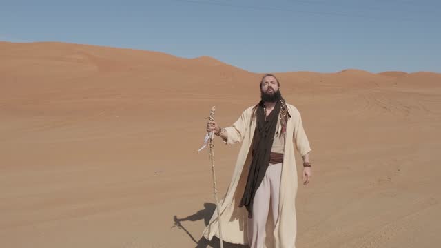 A young man in white shaman clothes with a staff in his hand walks on the sand of the desert against the backdrop of sunset