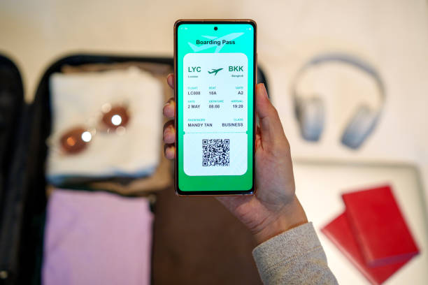 istock A close-up image shows a woman holding a smartphone displaying an electronic flight ticket while packing a suitcase with clothing, sunglasses, headphones, a laptop, and a passport. The suitcase is placed on a bed. 1490581435