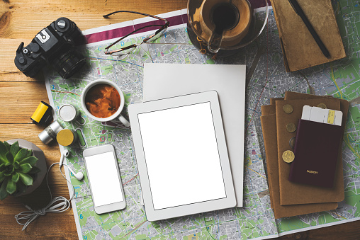 Travel concept flat lay table top shot on wooden background. Map, coffee, passport, glasses, earphones, succulent, pencils, notebook, camera and film rolls on the table with white cover magazine and blank screen digital tablet, smart phone.