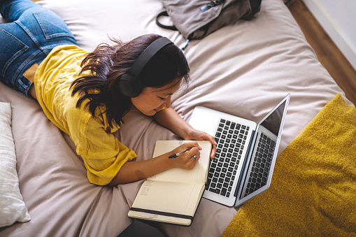 Top view of teenage asian college girl wearing headphones lying on bed planning week on paper planner. Doing homework, studying at home using laptop. Lifestyle concept.