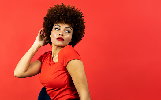 A dark-skinned girl with curly afro hair posing sitting on a red background in a red colored blouse. The woman touches her hair while looking back. Afro female fashion concept. Copyspace.