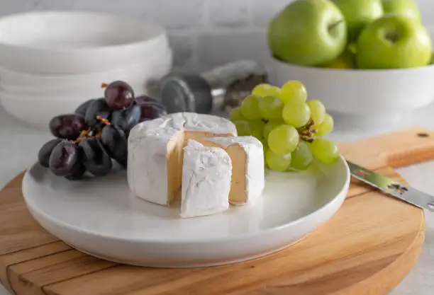 Round brie or soft cheese sliced on a plate with dark grapes and green grapes. Served ready to eat on kitchen table background on a wooden cutting board. closeup and front view